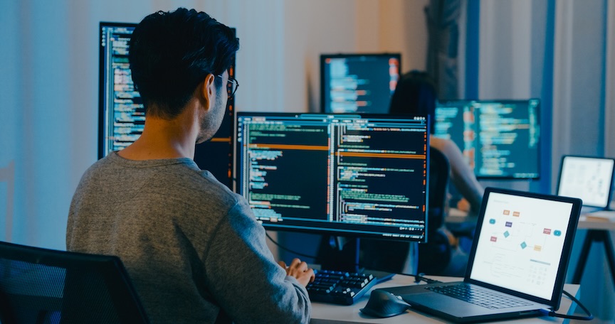 A developer at a multi-monitor workstation writing code, illustrating the evolution of software development from punch cards to AI-assisted coding